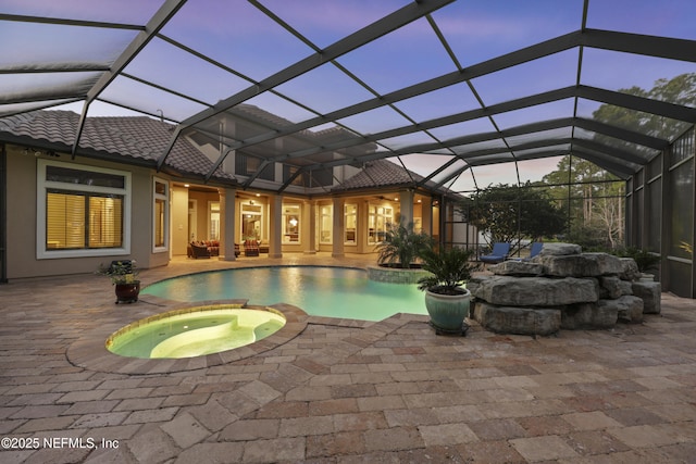 pool at dusk featuring a pool with connected hot tub, outdoor lounge area, a ceiling fan, glass enclosure, and a patio area