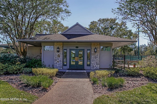 view of front facade featuring stone siding, french doors, roof with shingles, and fence