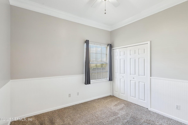 unfurnished bedroom featuring carpet, crown molding, a closet, a ceiling fan, and wainscoting