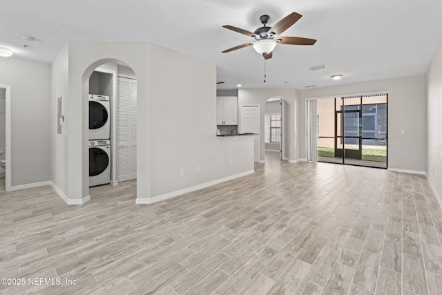 unfurnished living room featuring arched walkways, baseboards, light wood-style flooring, ceiling fan, and stacked washing maching and dryer