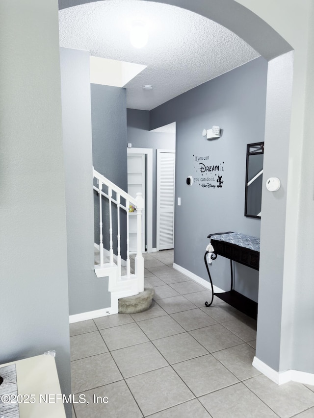 corridor with a textured ceiling, arched walkways, light tile patterned flooring, baseboards, and stairs