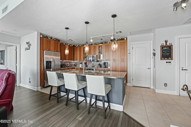 kitchen with stone counters, pendant lighting, appliances with stainless steel finishes, brown cabinetry, and an island with sink