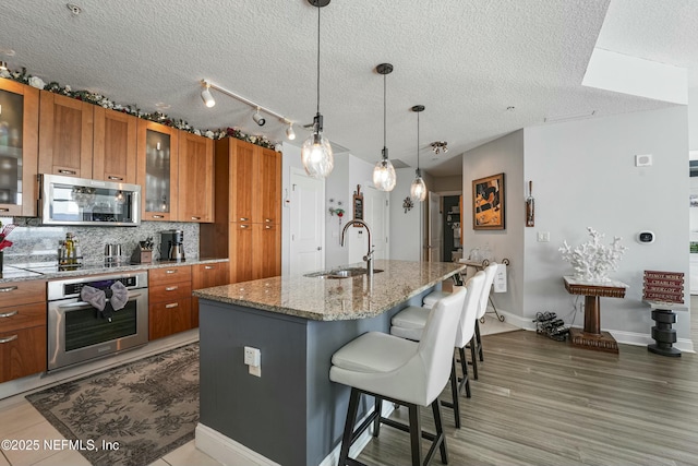 kitchen featuring a kitchen island with sink, a sink, appliances with stainless steel finishes, brown cabinets, and glass insert cabinets