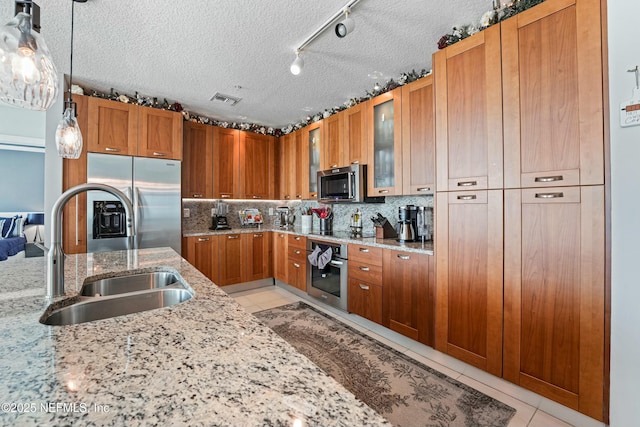 kitchen featuring light tile patterned floors, a sink, appliances with stainless steel finishes, brown cabinetry, and glass insert cabinets