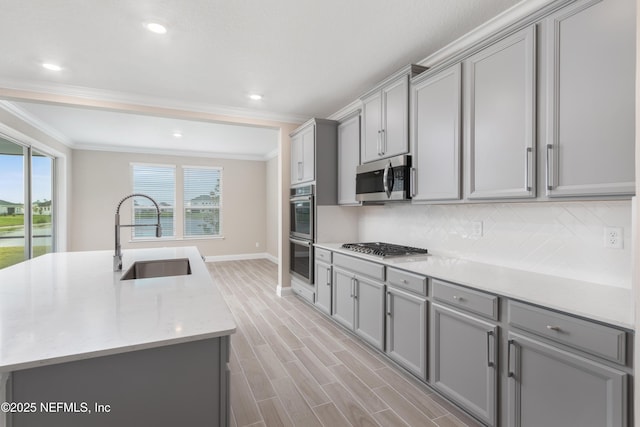 kitchen with a center island with sink, gray cabinetry, appliances with stainless steel finishes, a sink, and light stone countertops