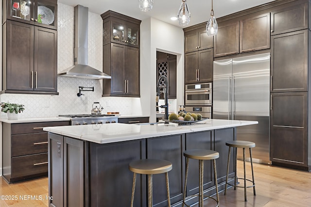 kitchen featuring dark brown cabinetry, wall chimney exhaust hood, appliances with stainless steel finishes, a breakfast bar, and light wood-type flooring