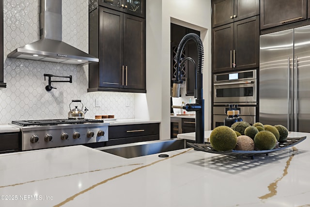 kitchen featuring built in fridge, dark brown cabinets, wall chimney range hood, decorative backsplash, and light stone countertops