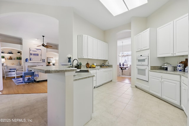 kitchen with a peninsula, arched walkways, a sink, white double oven, and ceiling fan with notable chandelier