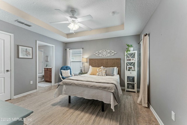 bedroom featuring a textured ceiling, ensuite bathroom, visible vents, baseboards, and a tray ceiling