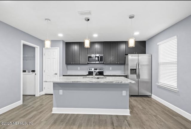 kitchen featuring stainless steel appliances, a kitchen island with sink, hanging light fixtures, and light stone counters