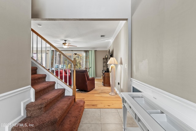 stairs featuring visible vents, wainscoting, ceiling fan, ornamental molding, and tile patterned flooring