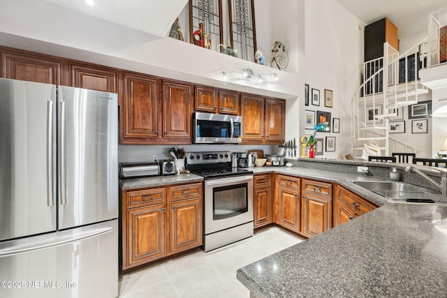 kitchen featuring brown cabinets, stainless steel appliances, dark countertops, a towering ceiling, and a sink