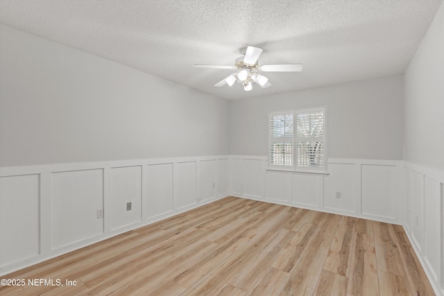 unfurnished room featuring a ceiling fan, a wainscoted wall, light wood-style flooring, and a textured ceiling