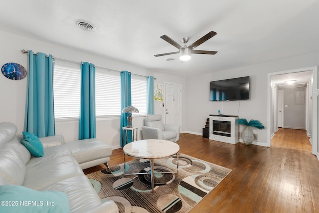 living room with dark wood finished floors, visible vents, a glass covered fireplace, ceiling fan, and baseboards