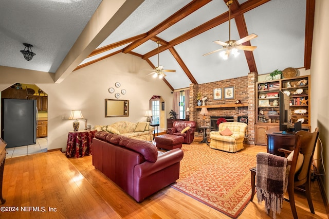 living room featuring high vaulted ceiling, light wood-style flooring, a fireplace, a ceiling fan, and beamed ceiling