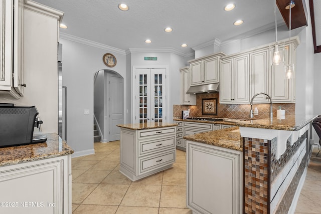 kitchen featuring backsplash, under cabinet range hood, arched walkways, cream cabinetry, and stone countertops