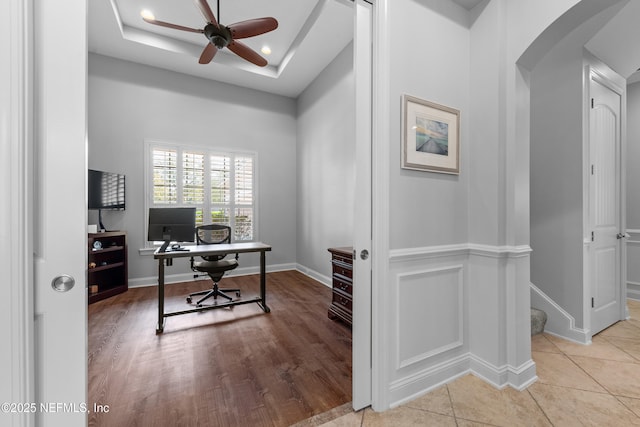 tiled home office featuring a wainscoted wall, arched walkways, a decorative wall, a raised ceiling, and a ceiling fan