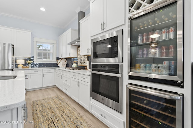 kitchen with wall chimney exhaust hood, appliances with stainless steel finishes, white cabinets, and ornamental molding