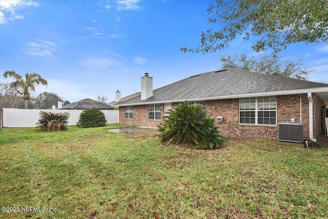 rear view of house with brick siding, a shingled roof, a lawn, central AC unit, and fence