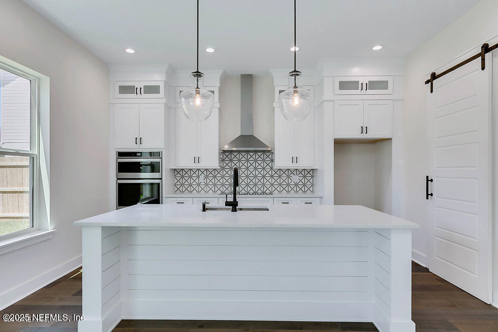 kitchen with double oven, a barn door, decorative backsplash, wall chimney exhaust hood, and a center island with sink