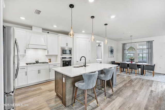 kitchen featuring a kitchen island with sink, stainless steel appliances, light countertops, white cabinetry, and a sink