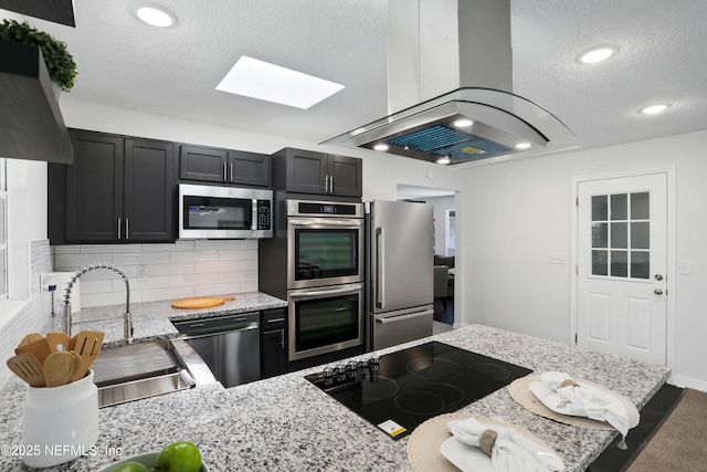 kitchen with island exhaust hood, stainless steel appliances, a sink, light stone countertops, and dark cabinets