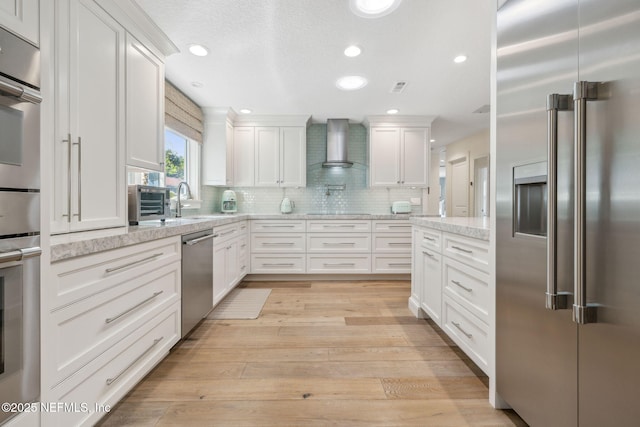 kitchen with white cabinets, appliances with stainless steel finishes, light wood-type flooring, wall chimney range hood, and backsplash