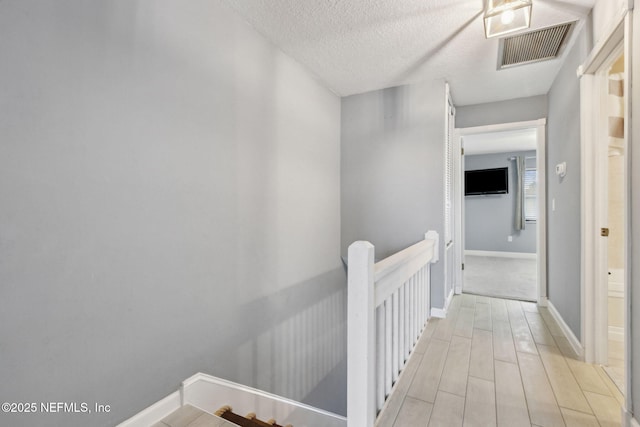 corridor featuring baseboards, visible vents, a textured ceiling, an upstairs landing, and light wood-style floors