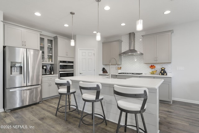 kitchen with a center island with sink, stainless steel appliances, light countertops, hanging light fixtures, and wall chimney exhaust hood