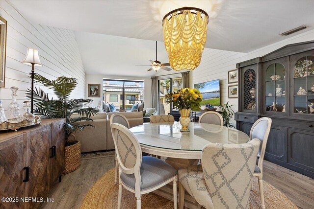 dining space featuring visible vents, wood walls, vaulted ceiling, ceiling fan with notable chandelier, and wood finished floors