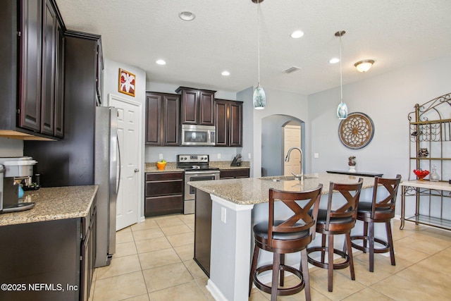 kitchen featuring arched walkways, light tile patterned flooring, stainless steel appliances, a sink, and a center island with sink