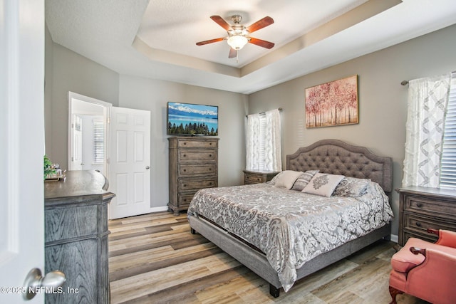 bedroom with a textured ceiling, light wood-style flooring, a ceiling fan, baseboards, and a tray ceiling