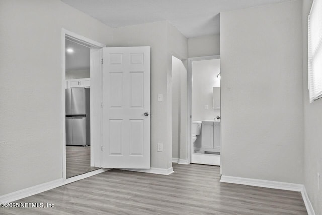 unfurnished bedroom featuring light wood-type flooring, freestanding refrigerator, baseboards, and ensuite bathroom