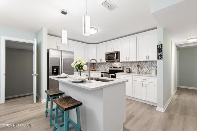 kitchen with decorative light fixtures, stainless steel appliances, white cabinets, a kitchen island with sink, and a sink