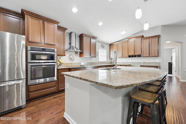 kitchen featuring an island with sink, dark wood-style floors, appliances with stainless steel finishes, wall chimney range hood, and a sink