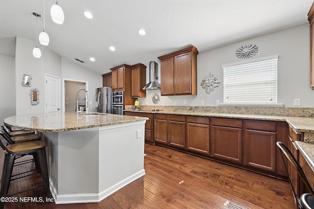 kitchen with dark wood finished floors, a kitchen island with sink, a sink, wall chimney range hood, and light stone countertops