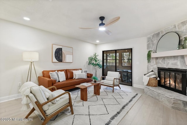 living area featuring light wood-style flooring, baseboards, a ceiling fan, and a stone fireplace