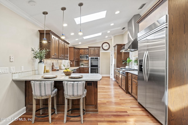 kitchen featuring light wood finished floors, decorative backsplash, wall chimney exhaust hood, built in appliances, and a peninsula