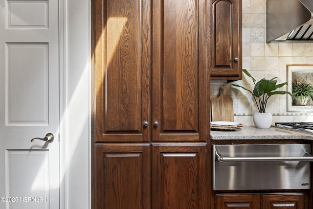 interior space featuring wall chimney range hood, dark brown cabinets, decorative backsplash, and a warming drawer