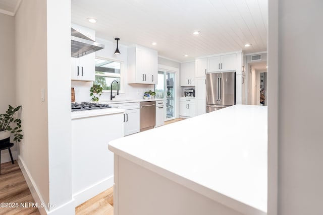 kitchen featuring pendant lighting, stainless steel appliances, light countertops, white cabinetry, and wall chimney range hood