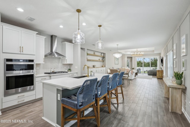 kitchen with a center island with sink, light countertops, appliances with stainless steel finishes, white cabinetry, and wall chimney exhaust hood