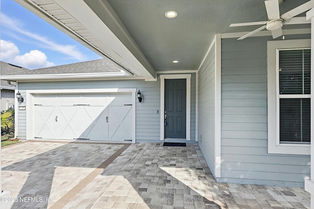 view of exterior entry featuring a garage, decorative driveway, roof with shingles, and ceiling fan