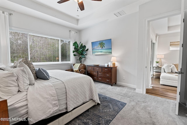 bedroom with a raised ceiling, light colored carpet, visible vents, ceiling fan, and baseboards