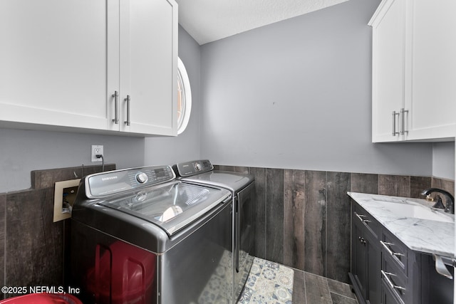 clothes washing area with a wainscoted wall, wood walls, a sink, cabinet space, and washing machine and clothes dryer