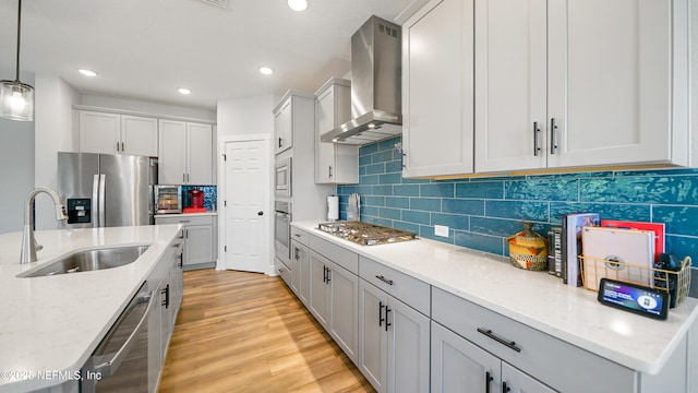 kitchen with light wood finished floors, stainless steel appliances, wall chimney range hood, pendant lighting, and a sink