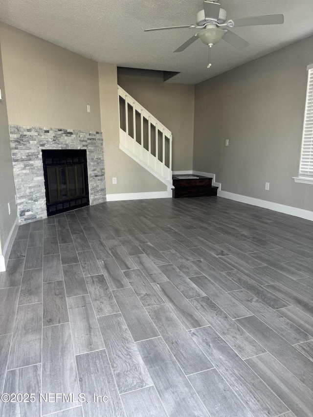 unfurnished living room with baseboards, ceiling fan, a stone fireplace, stairs, and wood finish floors