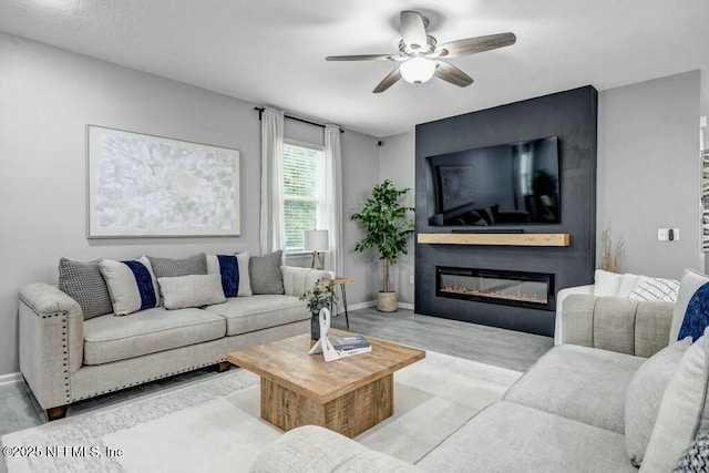 living area featuring a ceiling fan, a glass covered fireplace, baseboards, and wood finished floors