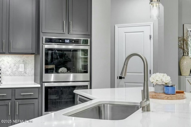 kitchen featuring stainless steel double oven, gray cabinetry, a sink, tasteful backsplash, and decorative light fixtures