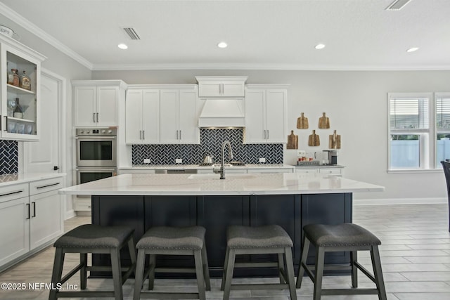 kitchen featuring appliances with stainless steel finishes, white cabinetry, an island with sink, and custom exhaust hood