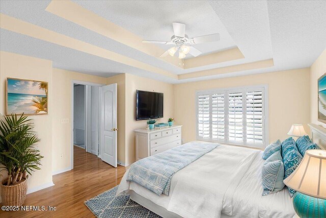 bedroom featuring a raised ceiling, ceiling fan, a textured ceiling, light wood-type flooring, and baseboards